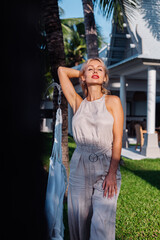 Outdoor portrait of rich caucasian woman in classic jumpsuit with red lipstick by hammock on vacation outside villa  hotel, sea side. 