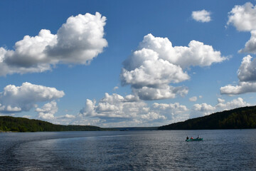 Fototapeta na wymiar Blue sky with beautiful white clouds over a wide river. Ripples on the surface of the water. Low cloud. Green forest on the banks. A small boat in the middle of the river.