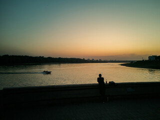 A man is standing on the embankment and watching a boat passing by in the sunset