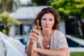 Young european woman smoking cigar lying on hammock outside tropical luxury villa hotel, sunset...