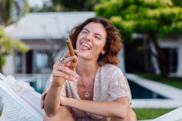 Fototapeta premium Young european woman smoking cigar lying on hammock outside tropical luxury villa hotel, sunset natural light. 