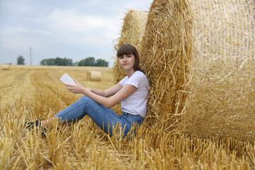 Naklejka na ściany i meble A young girl holds a phone in her hands,sitting on a harvested wheat field near large bales of straw.