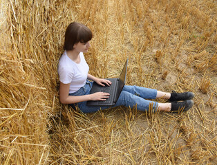 Naklejka na ściany i meble A young girl working on a computer, sitting on a field near the straw, top view.