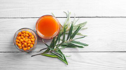 Sea buckthorn jam and fresh berries in glass jars and a green branch on a white wooden table