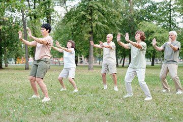 Focused coach raising hands forward while showing relaxing exercise to senior students at qigong practice - obrazy, fototapety, plakaty