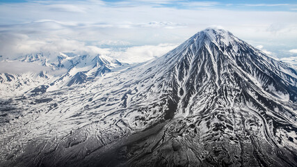 A black volcano with streaks of white snow on a background of mountain peaks and clouds. Kamchatka, a shot from an airplane.