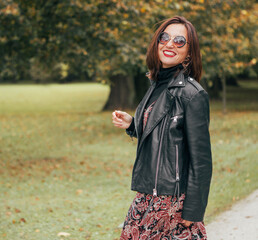 Smiling female dressed boho fashion style colorful long dress with black leather biker jacket in...