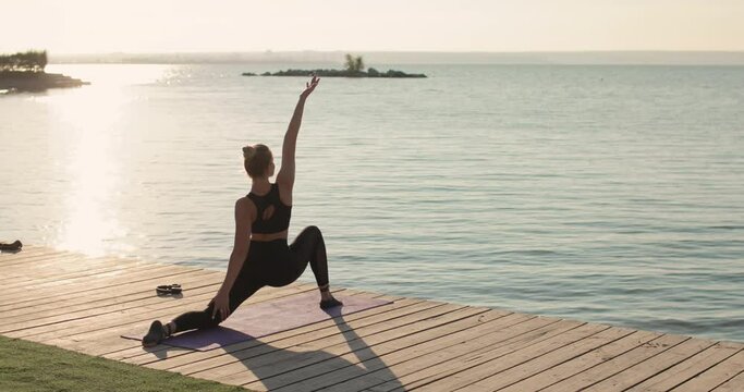 Young woman is training outdoors. Sports, yoga, stretch, wellness concept. Female is practicing yoga doing lunge warrior yoga pose on sport mat on river pier listening music in headphones, back view.