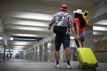 Couple stand with their backs and hold yellow suitcase by handle in underground passage. Man in shorts and plaid shirt point with his hand at timetable. Woman in shorts, T-shirt and white hat hold