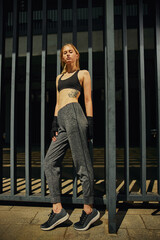 Young fit woman in sport suit stands by metal fence oudoors