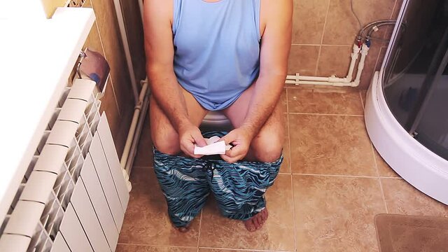 A man in shorts and glasses in the toilet sits on the toilet without a face