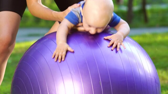 Young woman in sportswear doing exercises with baby on fitness ball in park. Mother holding little child by legs and swinging back and forth. Concept of gymnastics