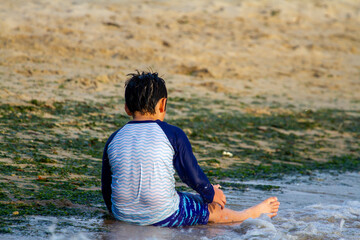 A young caucasian boy wearing rash guard long sleeve swimsuit is sitting on sand by the sea. He is...
