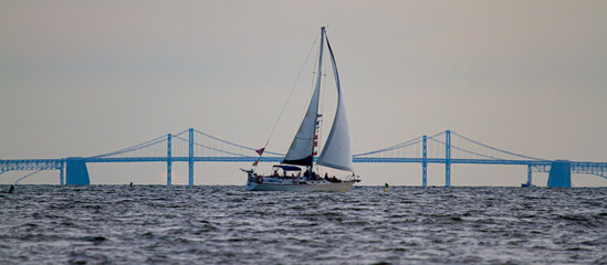 Panoramic image of a sailboat moving across Chesapeake Bay with the silhouette of the famous Bay Bridge in the background. There are people on the boat who are enjoying the sunset over the bay,.