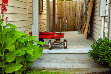 A red old rusty toy wagon was left on the doorway of an old house. The wooden porch the toy was left has grey paint which shows signs of aging. There are wooden fences on the other side of entrance. © Grandbrothers