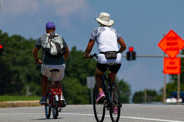 A senior couple wearing casual shorts and t shirts as well as hats is riding bikes near a highway on a sunny summer day. Man has a foldable pedal bike and the woman has a mountain bicycle.