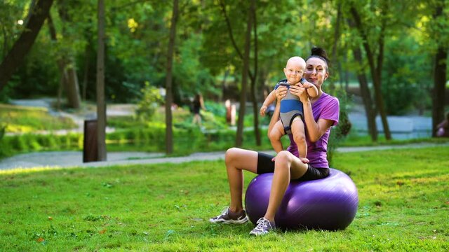 Sporty woman sitting on fitness ball and holding cute baby on hands, green park on background. Active mother doing gymnastics with little child outside. Concept of sport