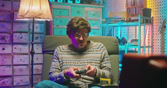 Caucasian young man nerd in glasses and with mustache sitting in front of vintage TV monitor and playing videogame with joystick. Male silly goofy play videogames. Retro style of 80's. Gamer from 90's