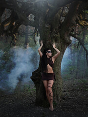 A beautiful wild Amazon girl with long legs stands in the smoke under a dry tree. Shaman girl with long black hair and ritual makeup.