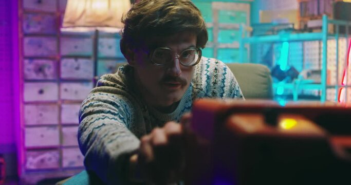 Caucasian funny young man nerd in glasses and with mustache sitting in retro style home and watching TV. Vintage television of 80's. Male standing and hitting side to catch signal. Goofy guy of 90's.