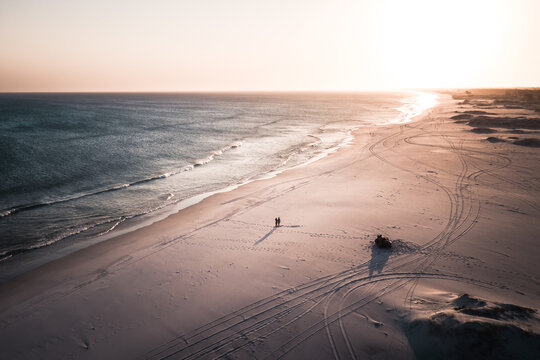 Aerial View Of Couple Standing Near Parked Vehicle On Empty Tropical Beach Marked With Tyre Tracks At Sunset, Arraial Do Cabo, Brazil