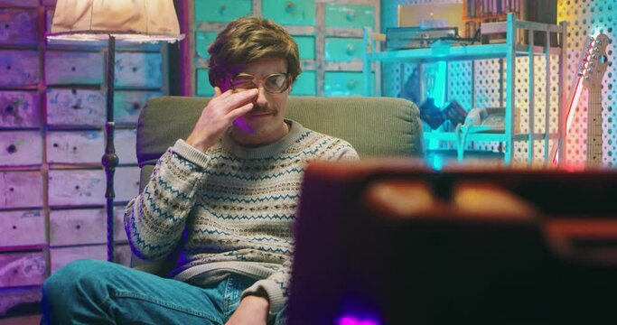 Caucasian funny young man nerd in glasses and with mustache sitting in retro style home and watching TV. Vintage television of 80's. Male crying at sad drama movie, film or program. Goofy guy of 90's.