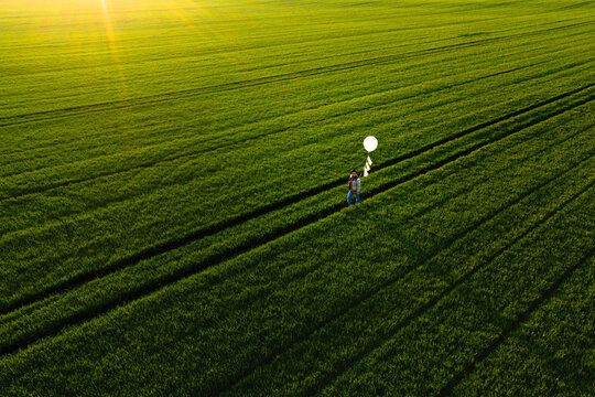 Aerial view of woman walking in green spring meadow while holding white balloon in her hands. Shot during golden hour at sunset near Kaunas, Lithuania.
