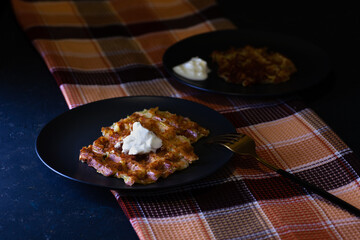 toasted Viennese zucchini waffles with sour cream sauce on two black plates, a checkered towel on a dark concrete background.