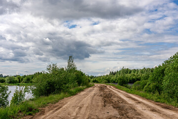 Fototapeta na wymiar Dirt road against blue sky, green forest and lake.