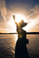 Samotna kobieta w kapeluszu na głowie nad brzegiem jeziora, ogląda zachód słońca.