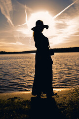 Samotna kobieta w kapeluszu na głowie nad brzegiem jeziora, ogląda zachód słońca.