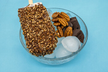 Walnut-dipped dark chocolate ice popsicle on a deep plate with ice and pieces of walnuts and chocolate on a blue background