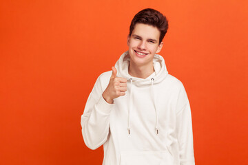 Positive young man in hoody with stylish hairdo showing thumds up and sincerely smiling. Well done! Indoor studio shot isolated on orange background