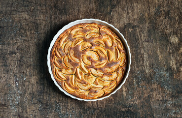 Traditional french homemade apple pie in a white ceramic baking form on old dark wooden background. Top view