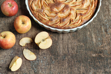 Traditional french homemade apple pie in a white ceramic baking form and autumn apples on old dark wooden background. Top view, copy space
