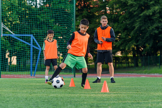 Persistent teen boy in sportswear trains football on soccer field and learns to circle the ball between training cones.