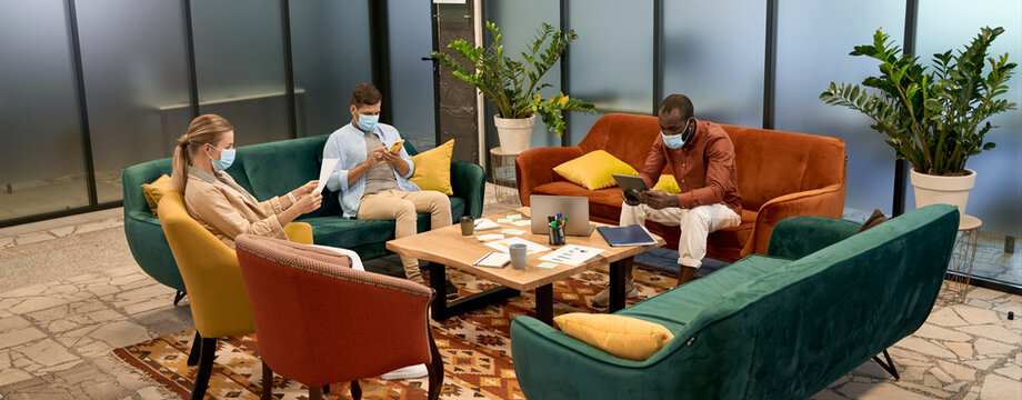 Keep Your Distance. Young People, Office Workers Wearing Medical Protective Masks Working Or Relaxing Together In The Modern Coworking Space. Social Distancing