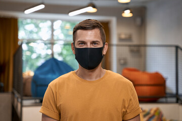 Working safely during coronavirus. Portrait of young man, male office worker wearing black protective mask and looking at camera, standing in the modern office