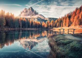 Lake with reflection in mountains at sunrise in autumn in Dolomites, Italy. Landscape with Antorno lake, small wooden bridge, trees with orange leaves, high rocks, blue sky in fall. Colorful forest