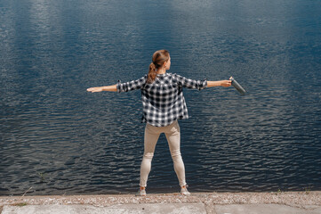 Young girl with her back in hat on the river bank, spread her hands in the sides, looks at the setting sun. Enjoys and meditates