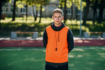 Young professional soccer coach in an orange vest and a whistle on neck at artificial football...