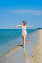 A young girl runs along the sandy sea beach along the surf. Splashes of sea water. The girl is wearing a white swimsuit and sunglasses. Freedom and carelessness. Outdoor fitness. Sunny day. Copy space