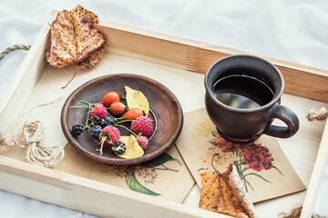 Fototapeta na wymiar Autumn cozy composition. A cup of coffee, autumn fruits and dried leaves on a wooden tray on a white bed. Morning coffee.
