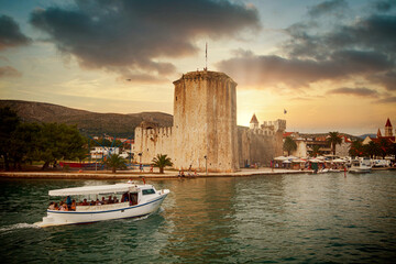 Touristic boat approaches Trogir harbor on the Dalmatian coast at sunset, with the tower of its...