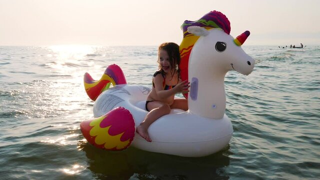 Three year old girl happily plays with her inflatable unicorn at the sea. The scene tells joy and happy childhood in the sunset lights.