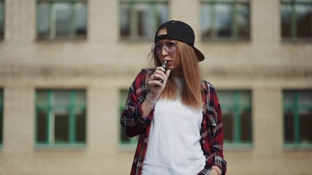 Pretty young hipster woman in black cap vape ecig. Girl vape popular ecig gadget, vaping device.Happy brunette vaper girl with e-cig. Portrait of smoker female model with electronic cigarette