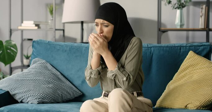 Young ill Arabian woman in hijab sitting on sofa at home and caughing as having coronavirus or flu. Sick muslim female in headscarf on couch in living room. Caugh and fever symptoms of coronavirus.