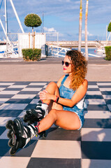 Young curly hair woman is sitting on the chess floor during wearing the roller skates