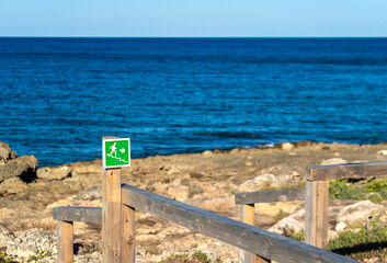Green safe exit sign with stylized running man, in a wooden walkway on a rocky cliff on the sea, in Posto Rosso, Marina of Alliste, Salento, Italy.