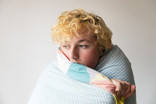A teenage male looks outside while wrapped in bedding comforter - unhappy at home alone